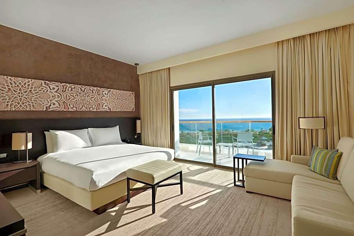 A hotel room with a large bed, a sofa, a small table, and a view of the ocean.