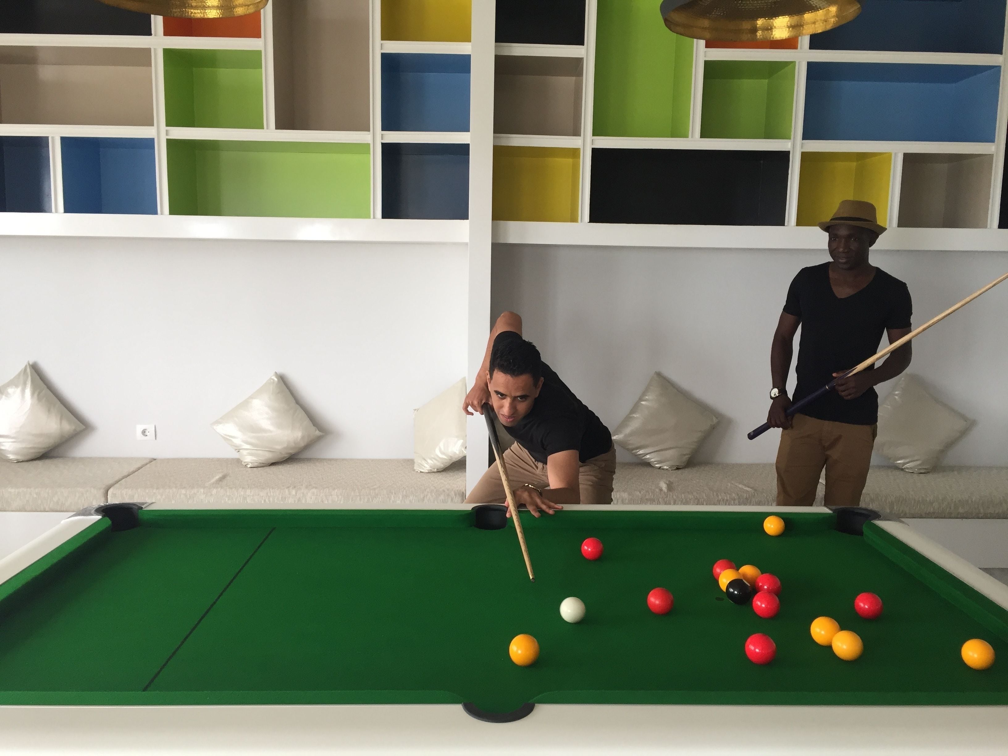 A pool table with balls and cues, a sofa with cushions, and a colorful shelving unit in the background.