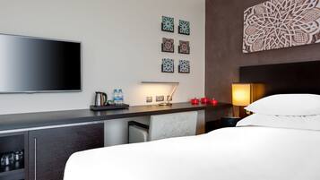 Camera d'albergo con TV a schermo piatto, una scrivania con lampada, una macchina del caffè e bottiglie d'acqua. Delle piastrelle decorative adornano la parete e un letto con biancheria bianca.