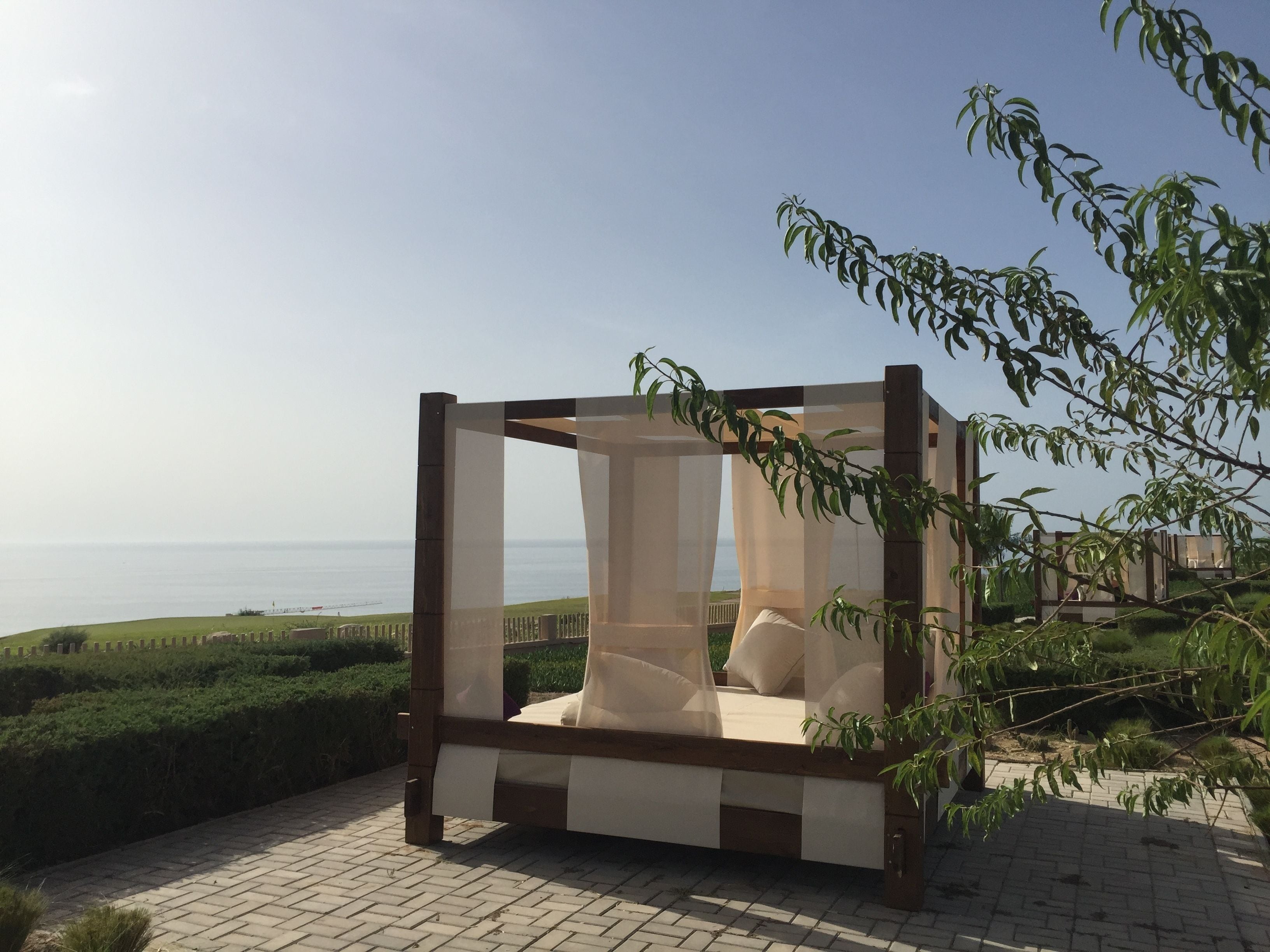 Ein hölzernes Baldachinbett mit Blick auf das Meer, umgeben von Grünpflanzen und einem gepflasterten Weg.