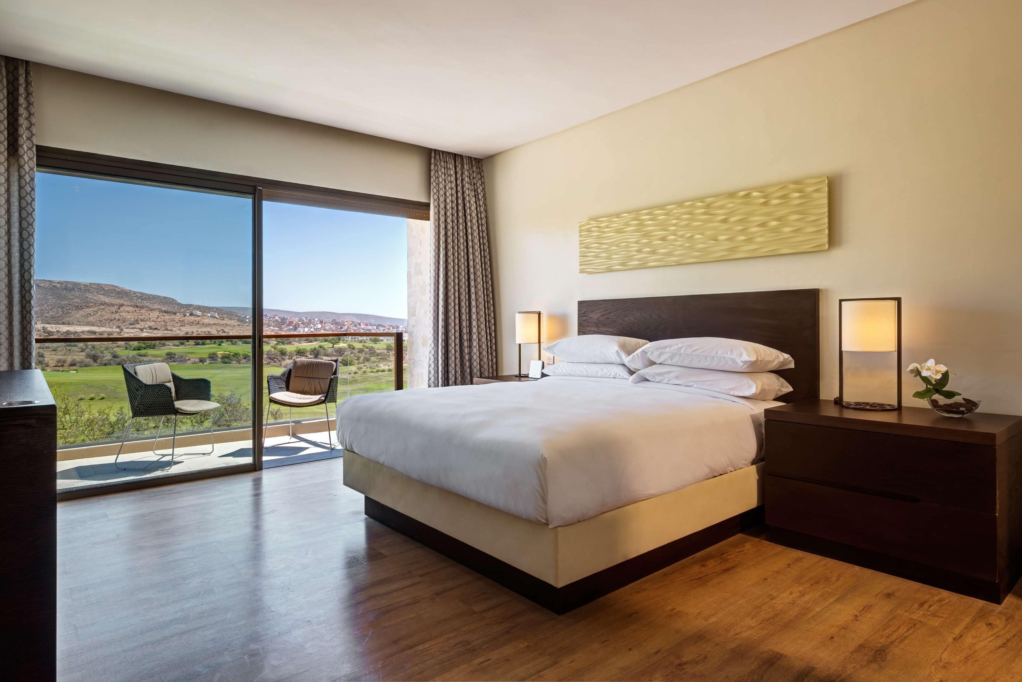 A modern hotel room with a large bed, a nightstand, a chair, and a view of a green landscape through a large window.