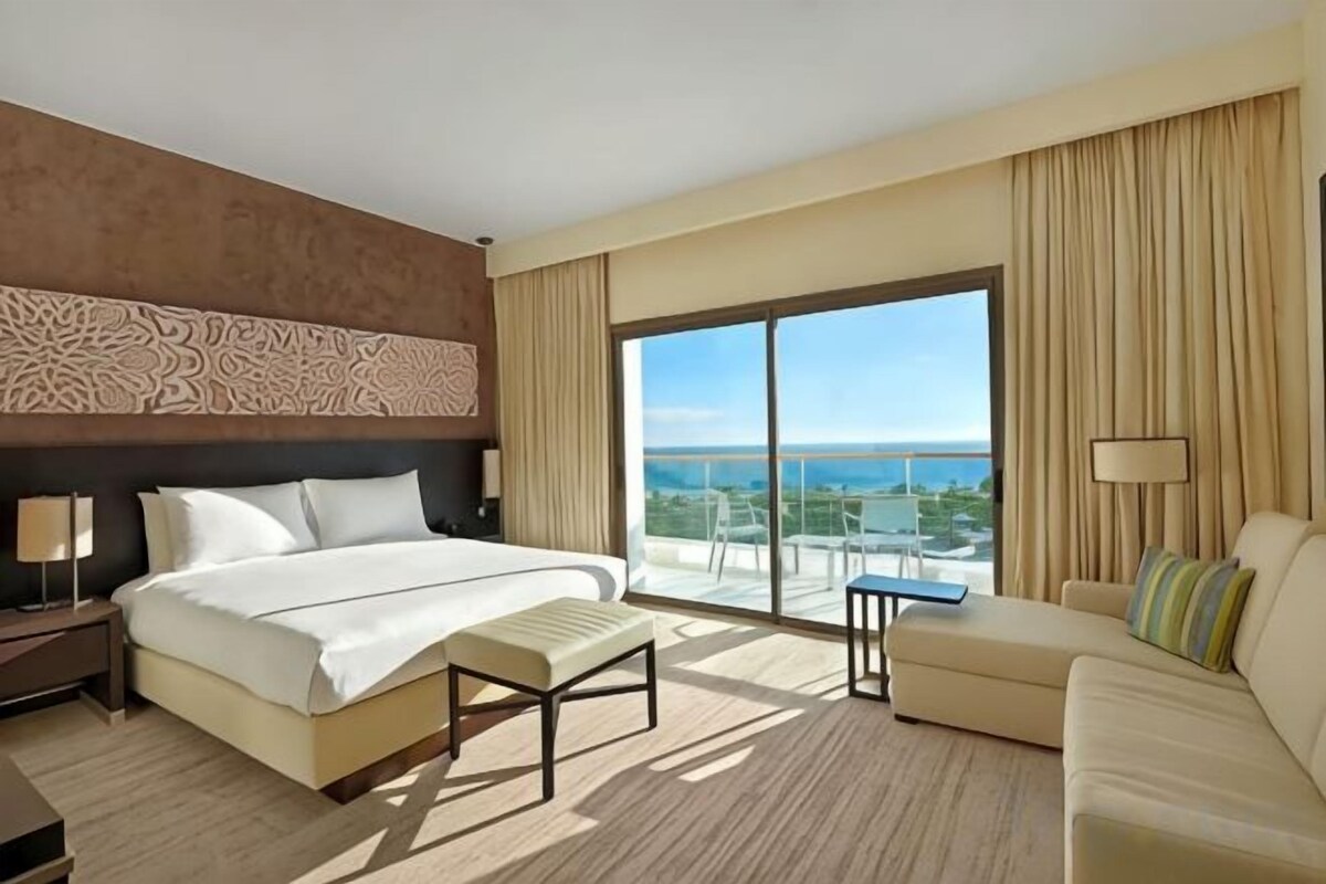 A hotel room with a large bed, a sofa, a small table, and a view of the ocean.