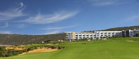 A modern hotel with a flat roof and multiple floors, situated on a green hill with a clear sky.