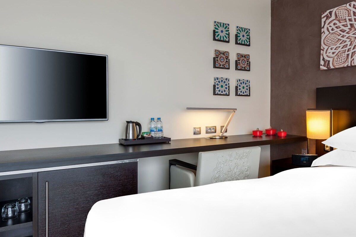 A hotel room with a bed, a TV, a desk with a lamp, a coffee maker, and decorative items.