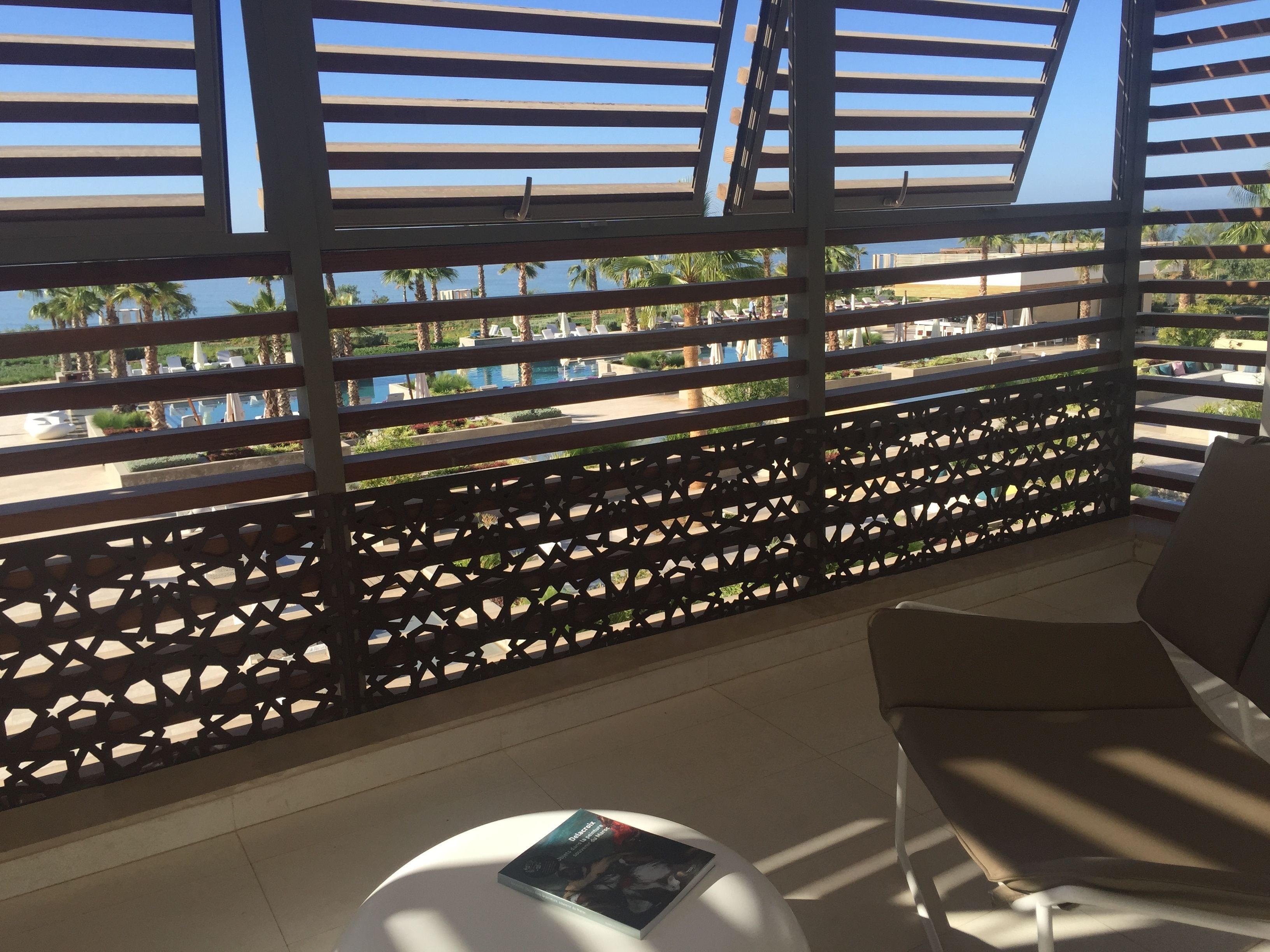 A balcony with a view of a pool and palm trees, a chair, and a small table with a magazine.