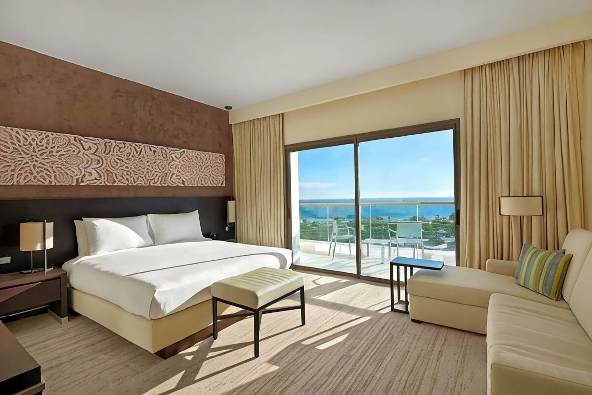 A hotel room with a large bed, a sofa, a small table, and a view of the ocean.