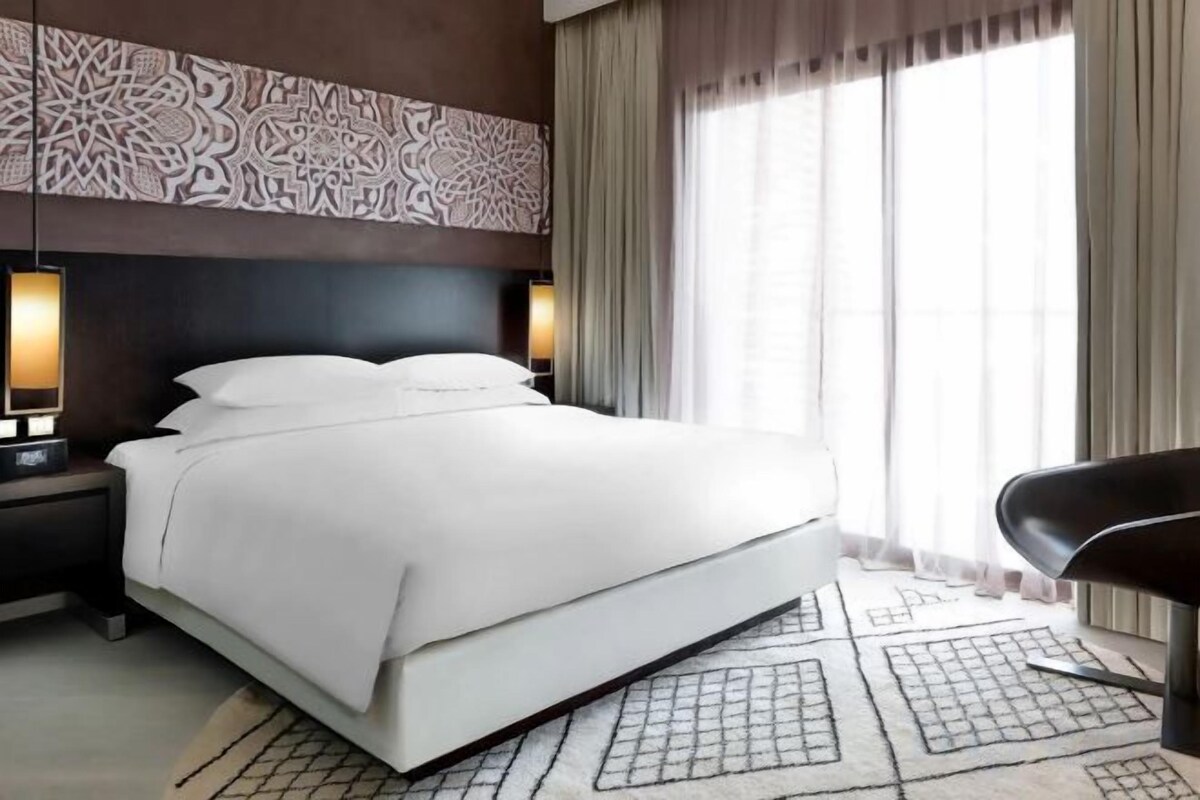 A hotel room with a large bed, a nightstand, a chair, and a window with curtains.