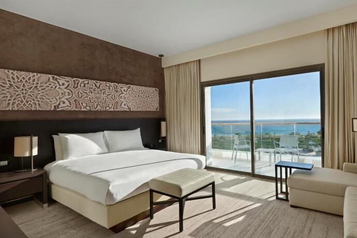 A hotel room with a large bed, a sofa, a balcony with a view, and a decorative wall panel.