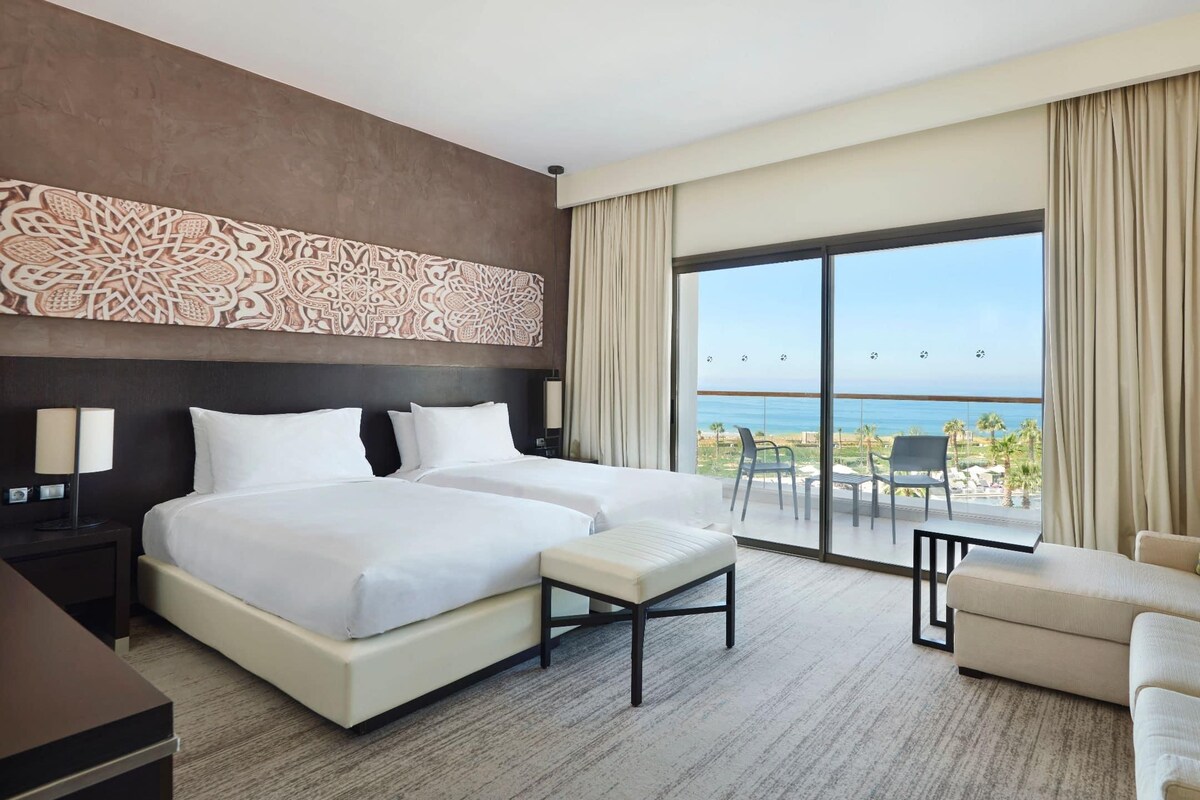 A hotel room with two beds, a sofa, a small table, and a balcony with a view of the sea.