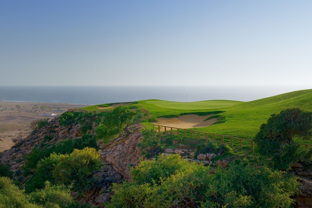 A golf course with a sandy bunker, a wooden fence, and a flagstick.