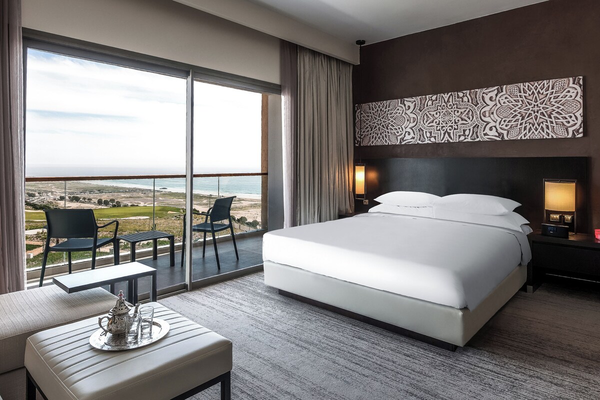 A hotel room with a large bed, a view of the beach, a balcony with chairs, and a decorative wall piece.