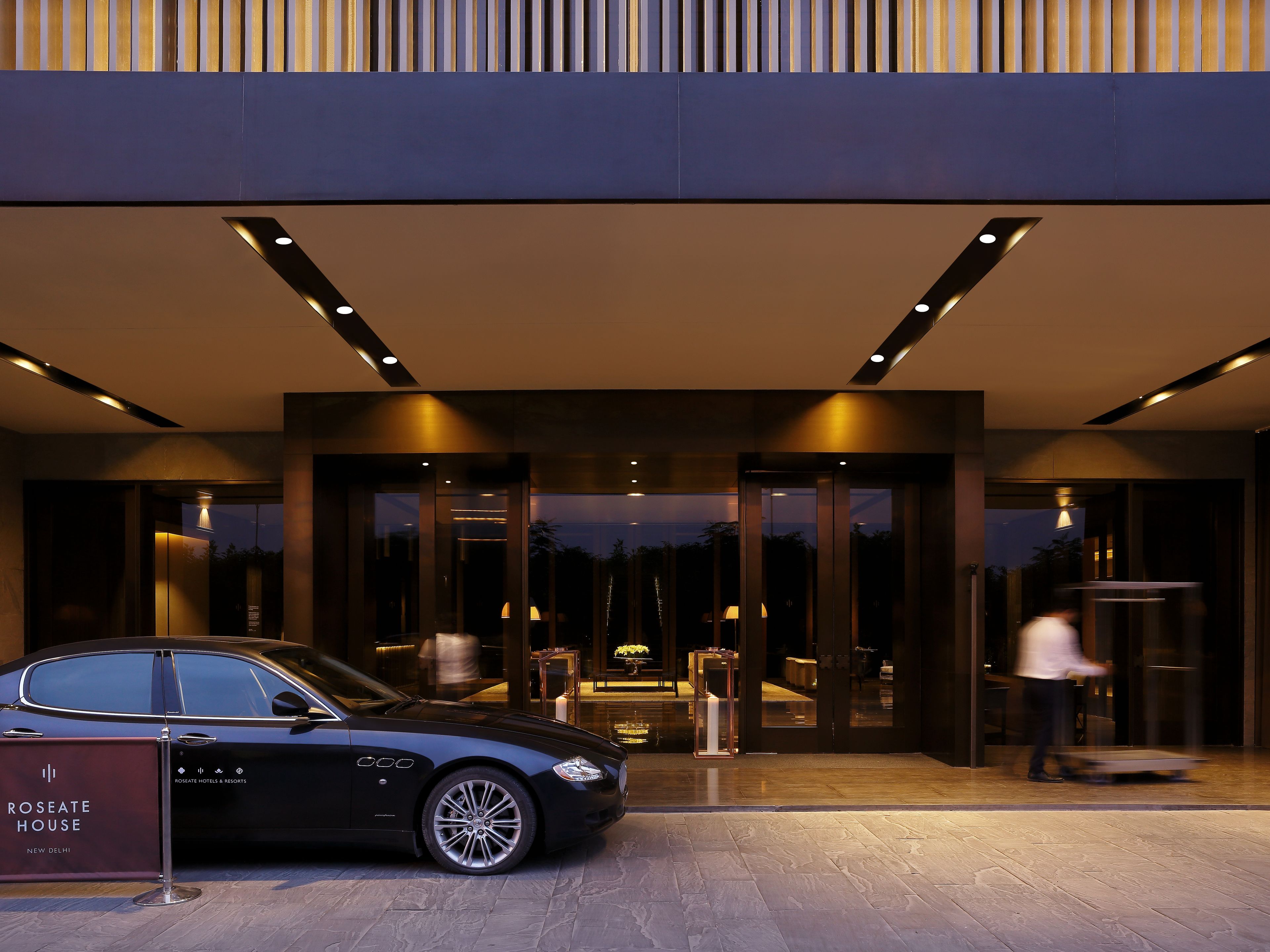 Entrada de un hotel moderno con un coche negro estacionado adelante, una puerta de cristal y una persona entrando.