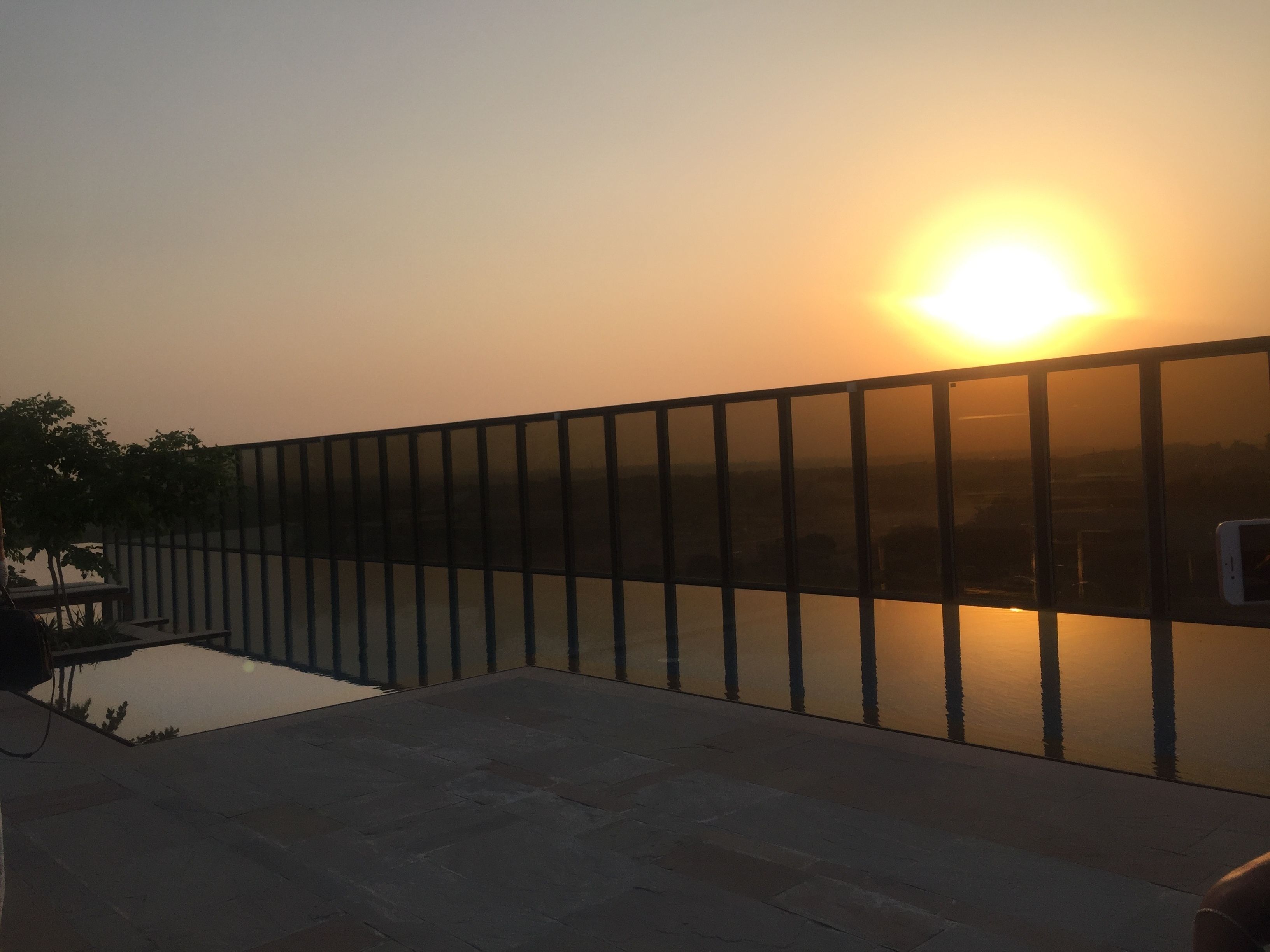 A sunset view from a balcony with a glass railing overlooking a body of water and a distant cityscape.