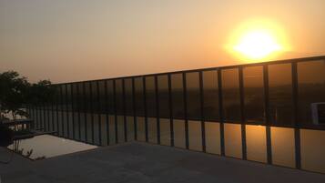 A sunset view from a balcony with a glass railing overlooking a body of water and a distant cityscape.