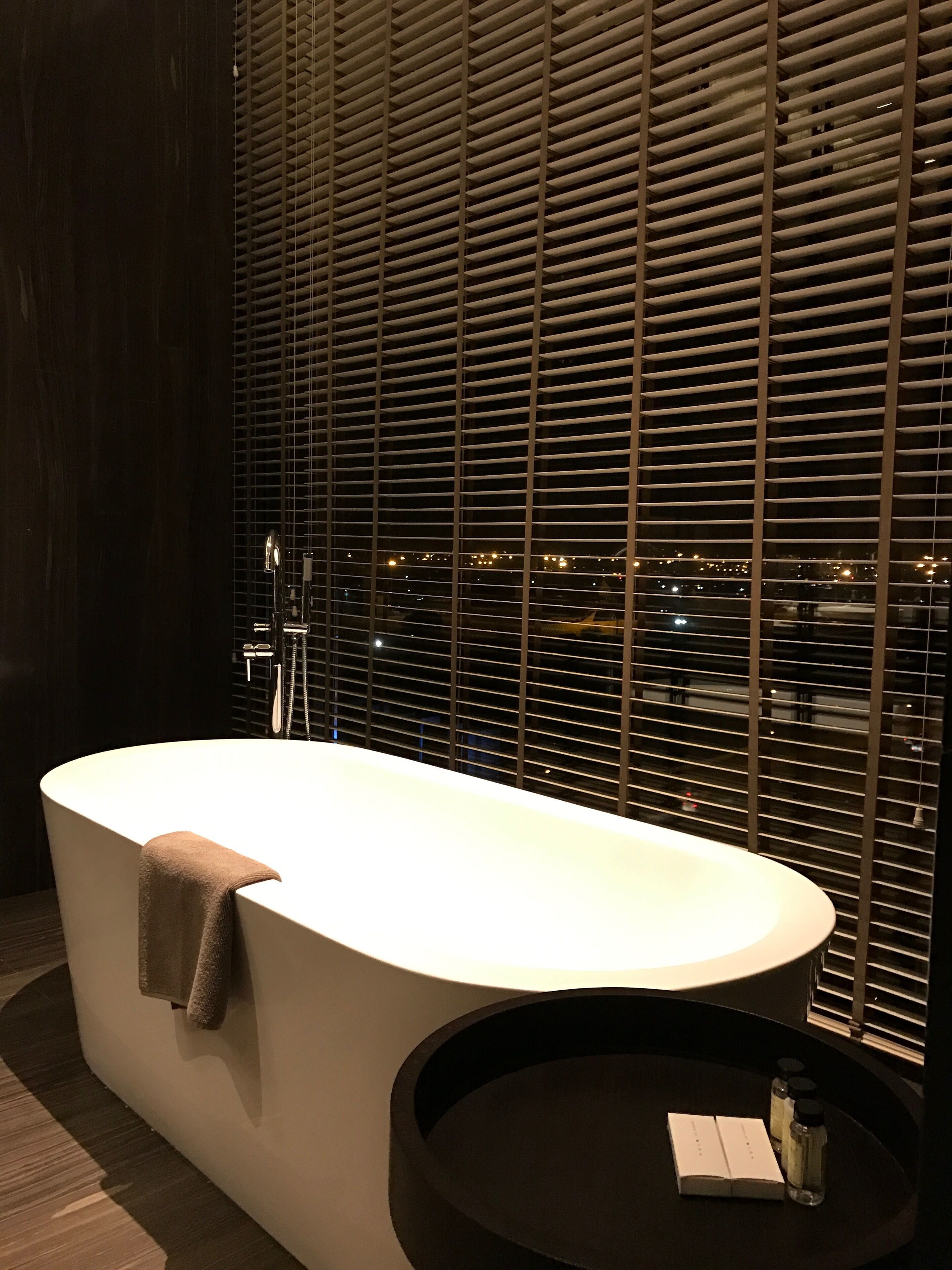 Une salle de bain moderne avec une baignoire sur pieds, une table ronde avec un livre et des bouteilles, et une vue sur la ville à travers de grandes fenêtres.
