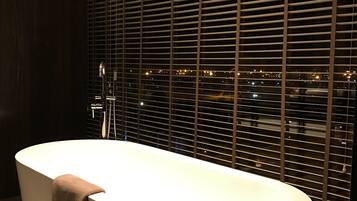 Une salle de bain moderne avec une baignoire sur pieds, une table ronde avec un livre et des bouteilles, et une vue sur la ville à travers de grandes fenêtres.