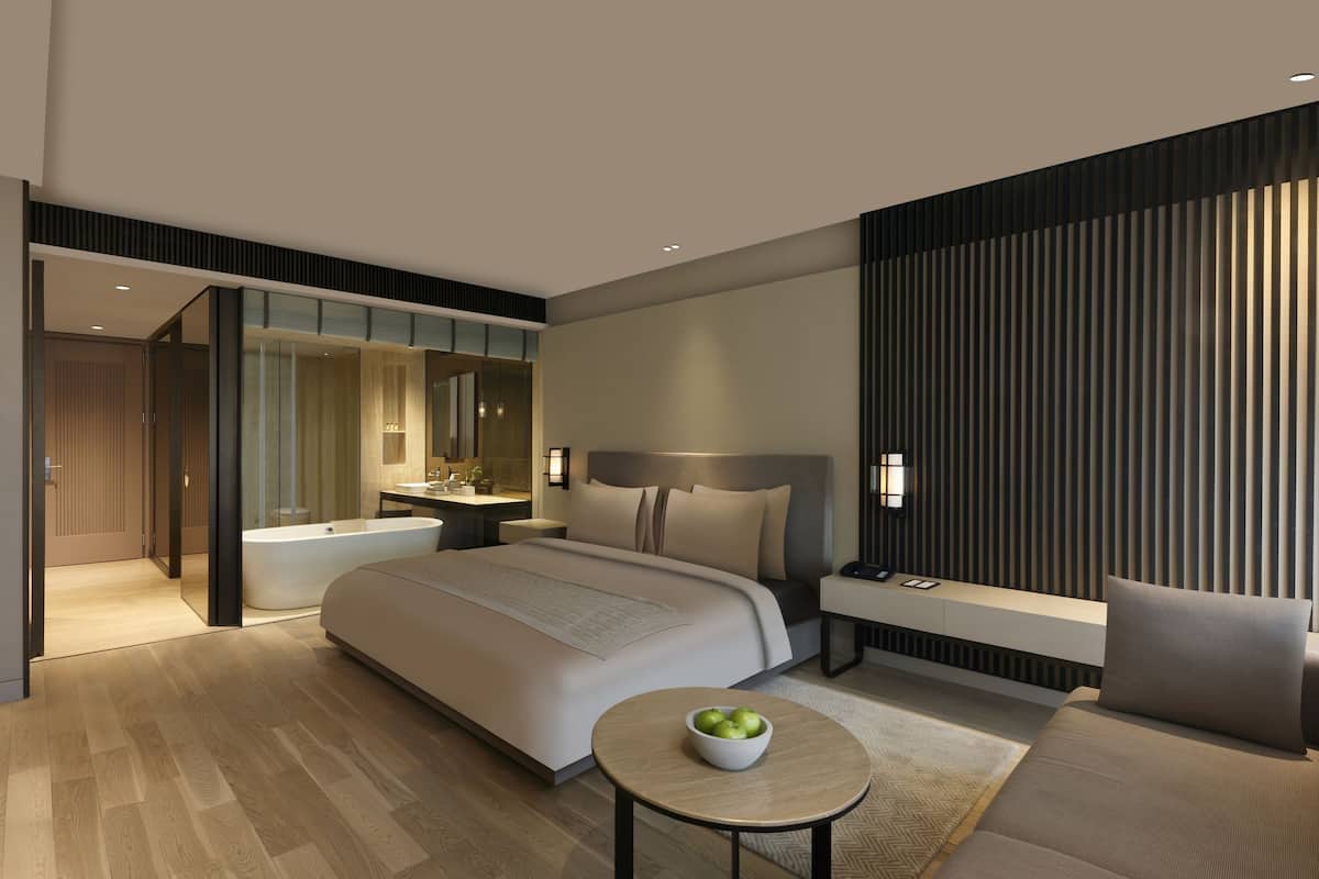 A modern hotel room with a large bed, a sitting area with a sofa and armchair, a small round table with a bowl of fruit, and a bathroom visible through an open door.