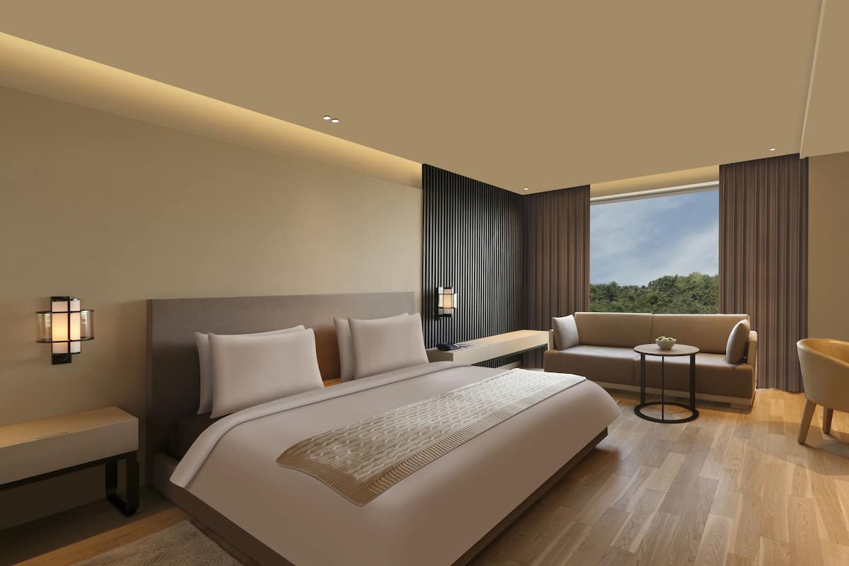 A modern hotel room with a large bed, a sofa, a desk, and a chair. There is a window with curtains, a nightstand, and a lamp.