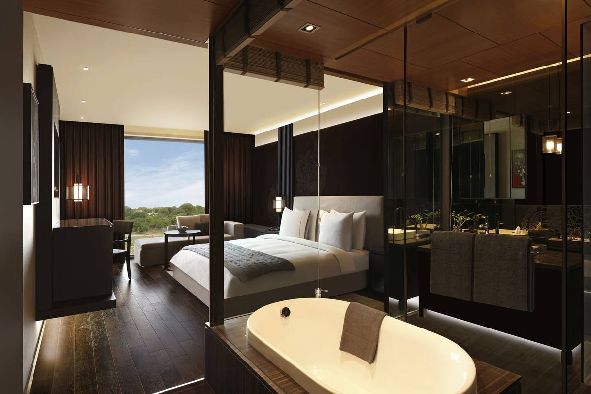 A modern hotel room with a large bed, a sitting area, a bathtub, and a view of the outdoors.