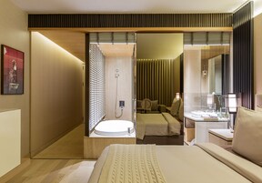 A modern hotel room with a large bed, a separate bathroom with a shower, and a small sitting area with a table and chair.