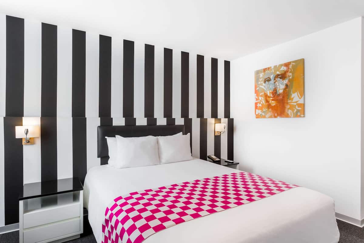 Una habitación de hotel con una cama, una mesita de noche, una lámpara de pared y un cuadro.