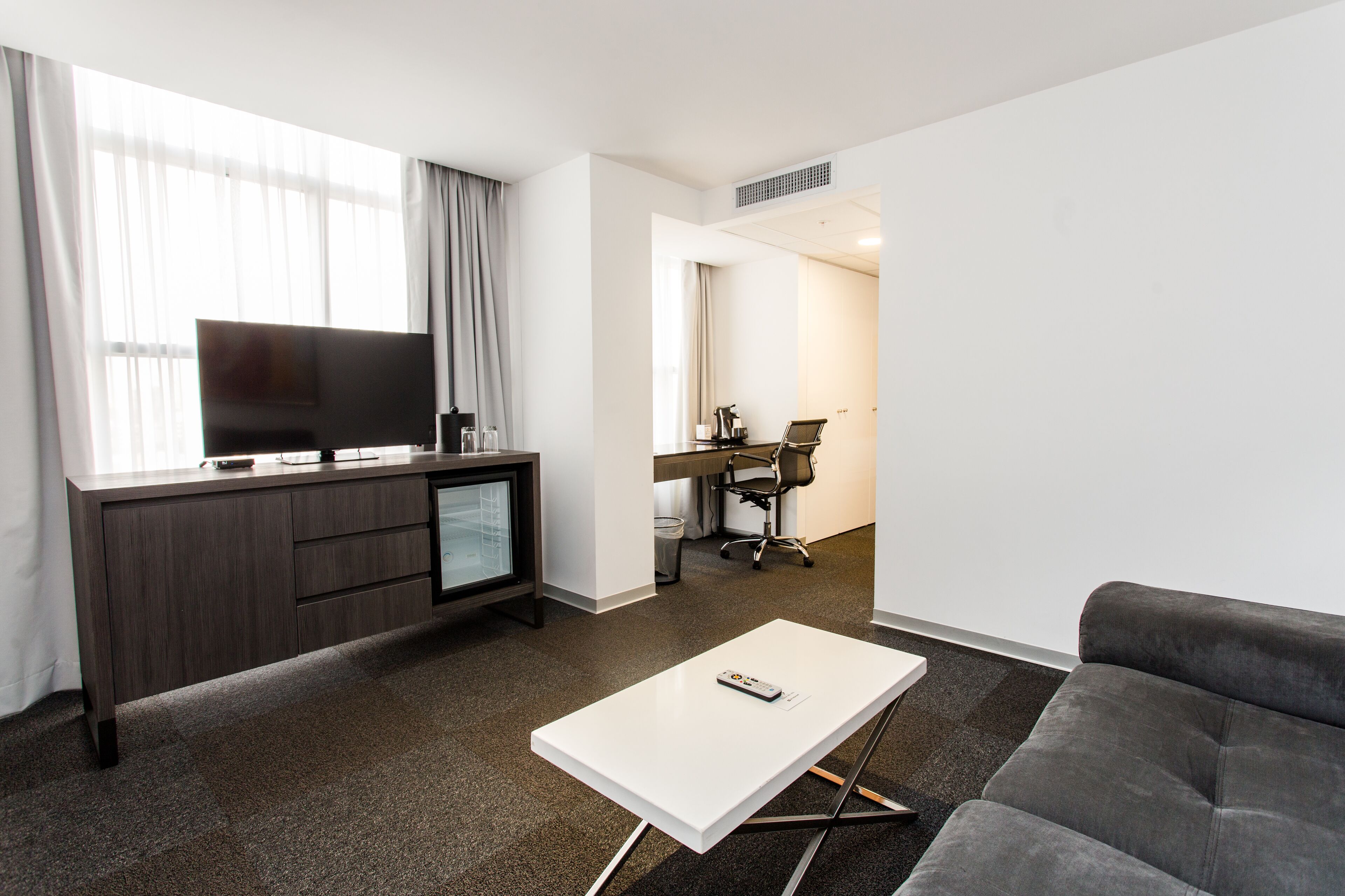 A modern hotel room with a flat-screen TV, a desk with a chair, a grey sofa, and a white coffee table.
