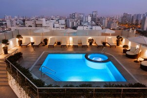 Piscina en la azotea con sillones de descanso y sombrillas, rodeada de vistas a la ciudad.