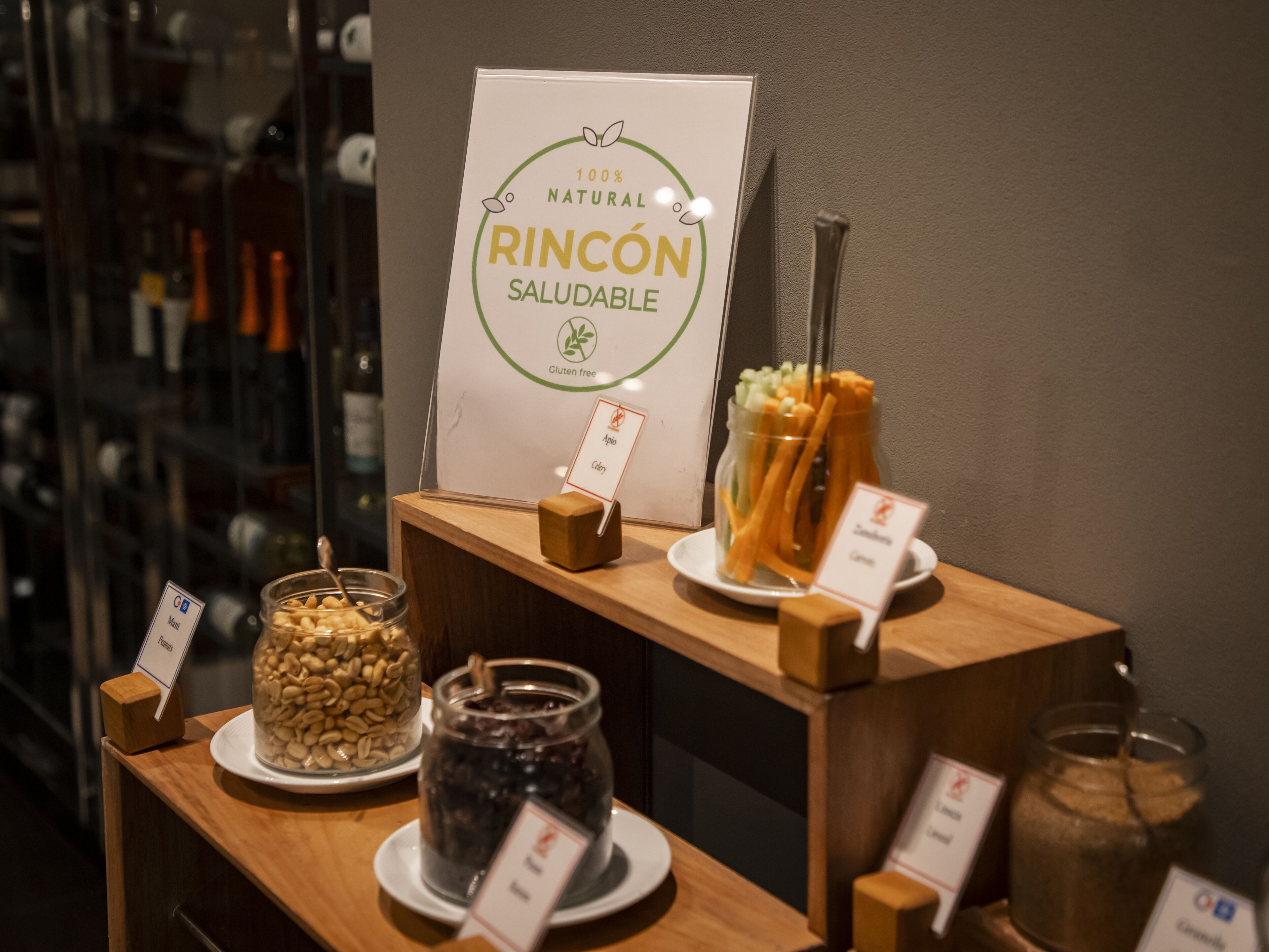 Un bar de aperitivos con opciones sin gluten, incluyendo frutos secos y palitos de zanahoria, y un cartel que indica que los aperitivos son 100% naturales.