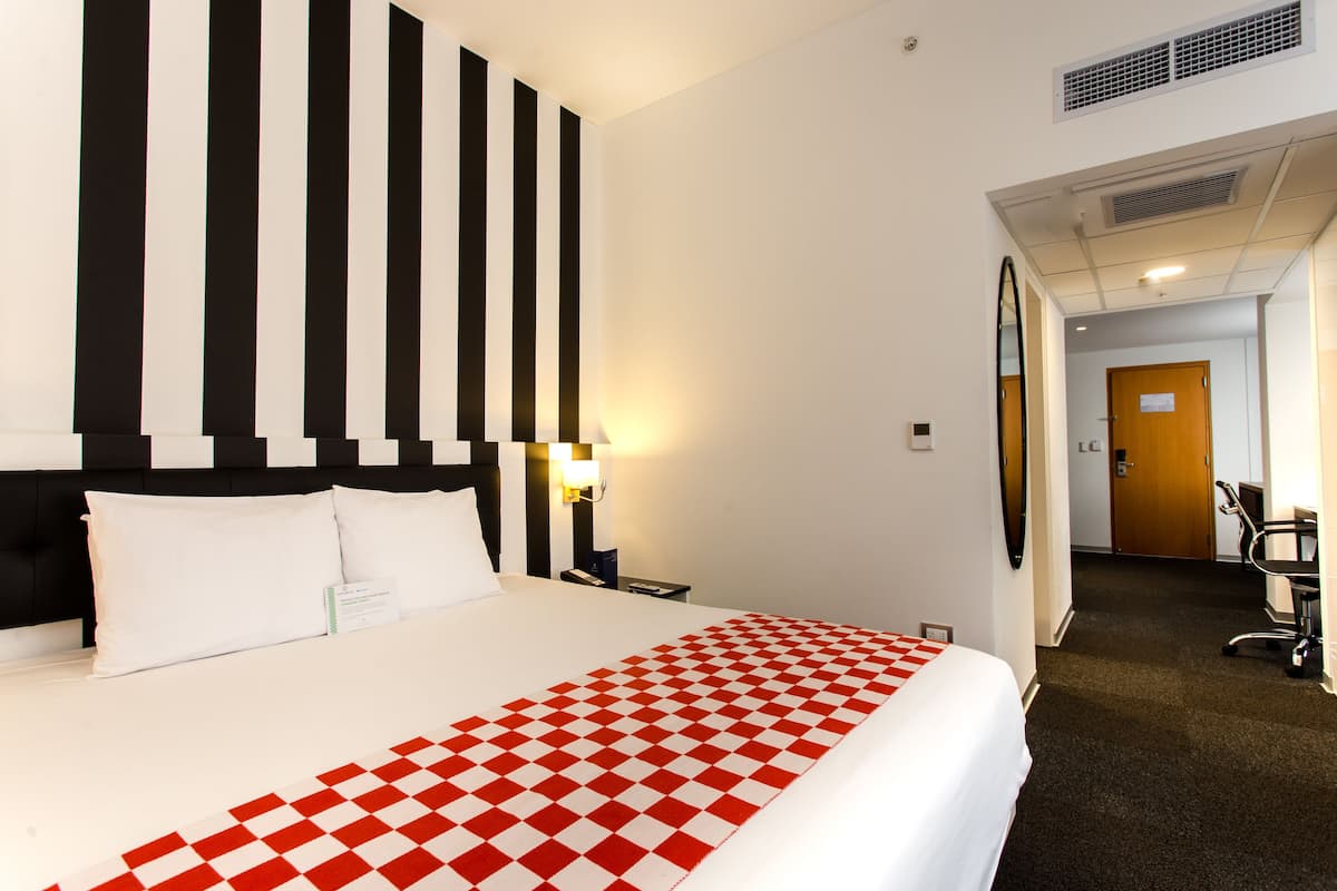 Habitación de hotel con una cama grande, cabecera a rayas blancas y negras, colcha a cuadros rojos y blancos, mesita de noche con lámpara y una puerta que da a un pasillo.