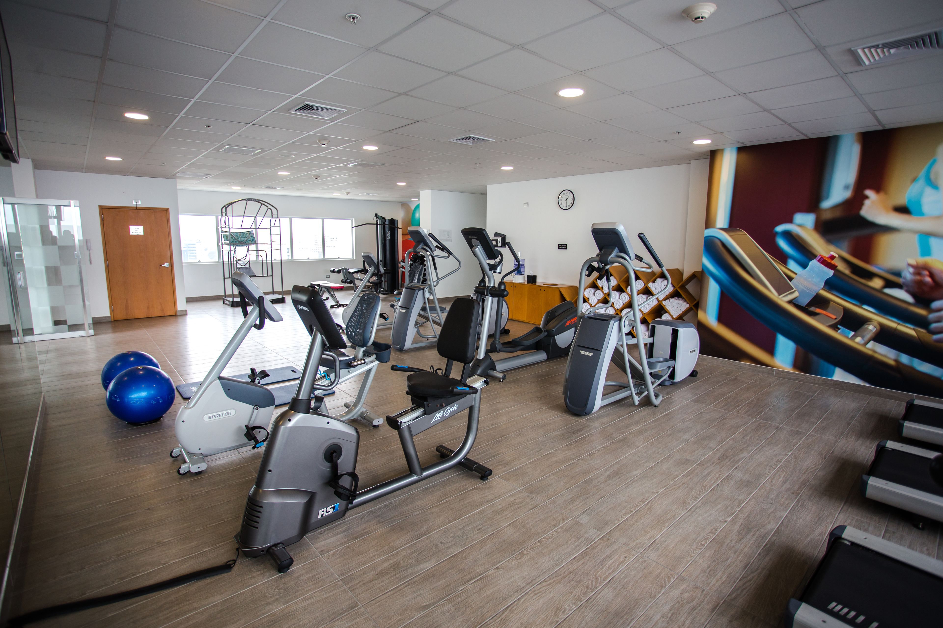 A gym with various exercise machines, a clock on the wall, and a large window in the background.