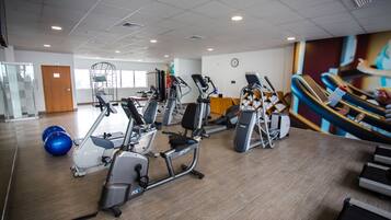 Un gimnasio con diversas máquinas de ejercicio, un reloj en la pared y un amplio ventanal al fondo.