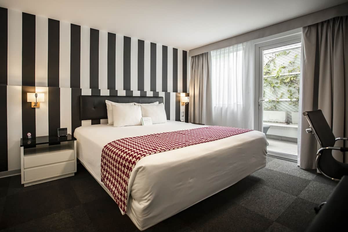 Habitación de hotel con una cama grande, pared a rayas, dos mesitas de noche con lámparas, una silla y vistas a zonas verdes.