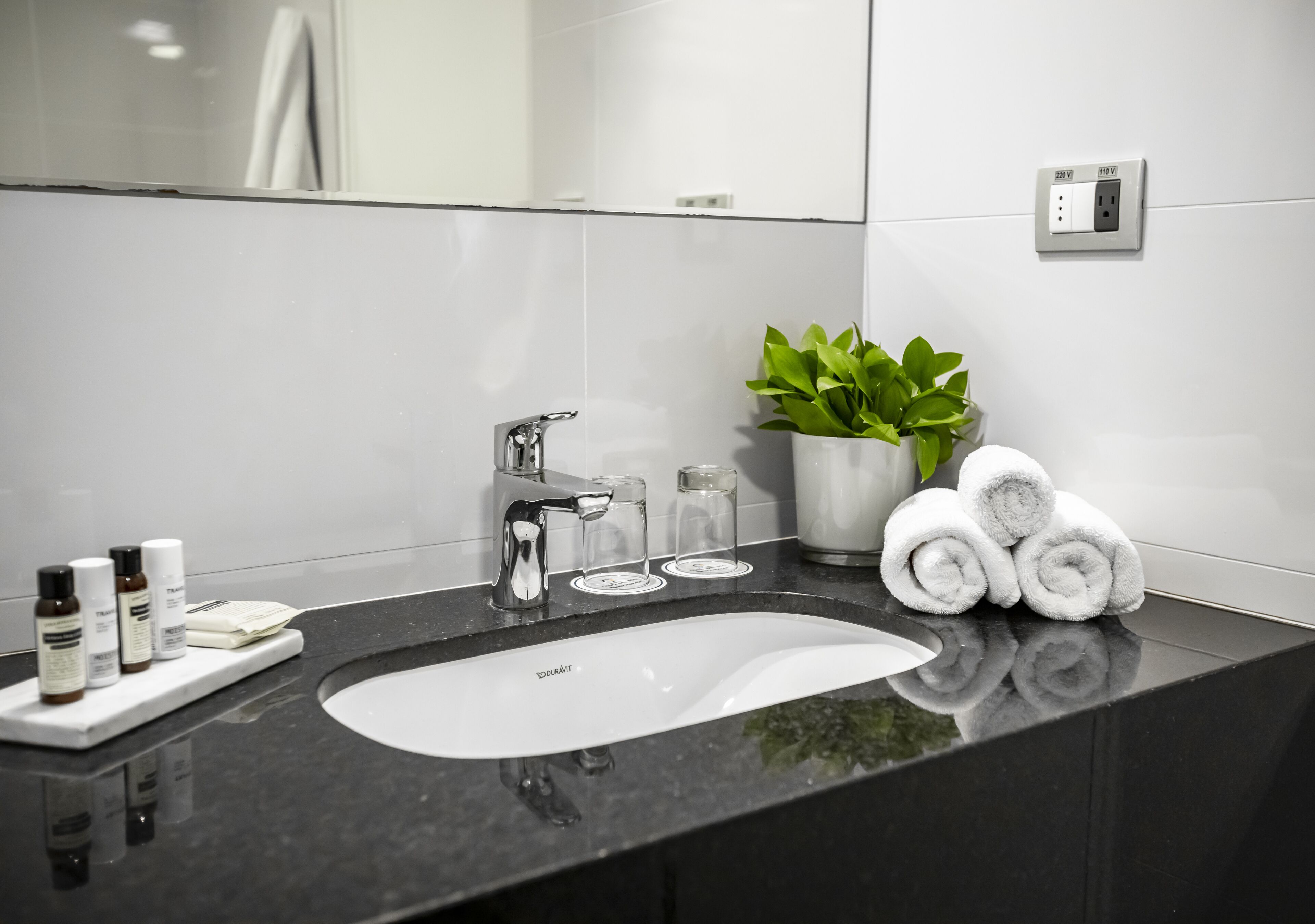 A modern bathroom with a black countertop, a white sink, a mirror, a plant, and neatly rolled towels.