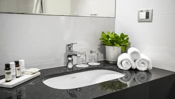 A modern bathroom with a black countertop, a white sink, a mirror, a plant, and neatly rolled towels.