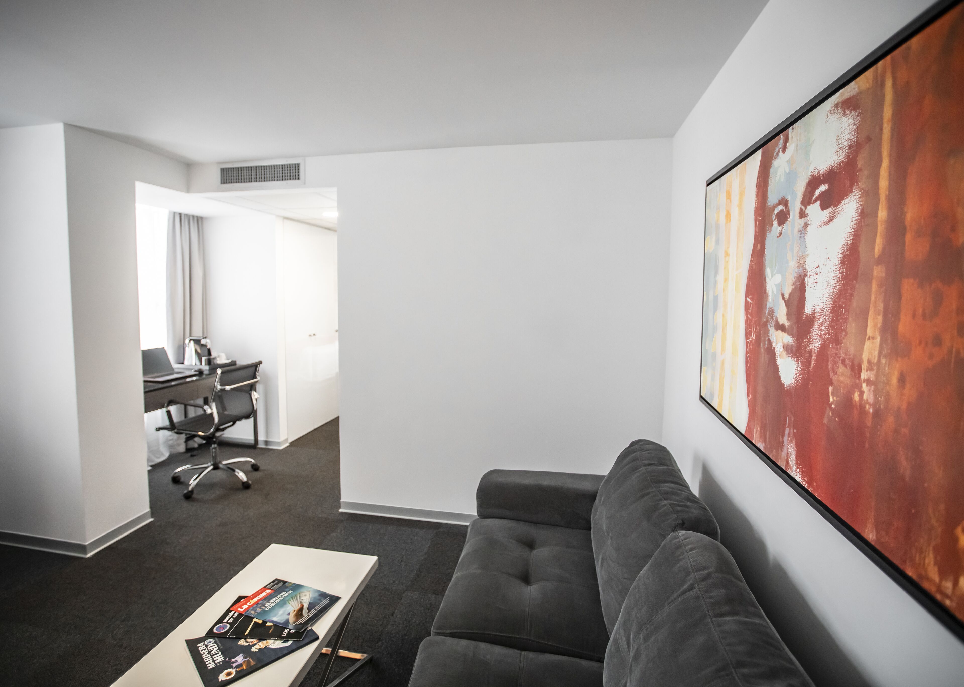 A modern hotel room with a grey sofa, a coffee table with magazines, a desk with a laptop, and a large abstract painting on the wall.