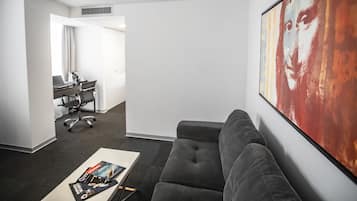 Una habitación de hotel moderna con un sofá gris, una mesa de centro con revistas, un escritorio con una computadora portátil y un cuadro abstracto grande en la pared.