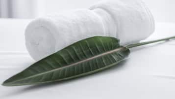 Una toalla blanca enrollada con cuidado y una hoja verde sobre una cama blanca.