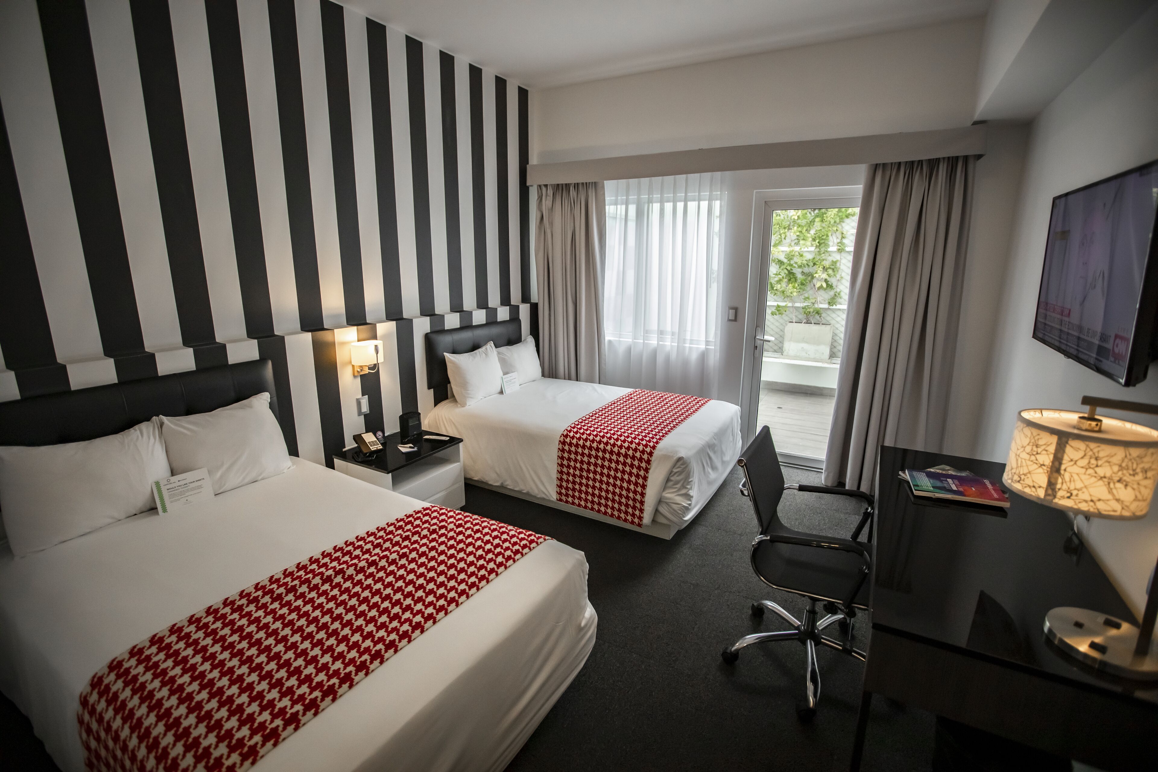Habitación de hotel con dos camas, un escritorio, una silla, un televisor y un balcón con cortinas.