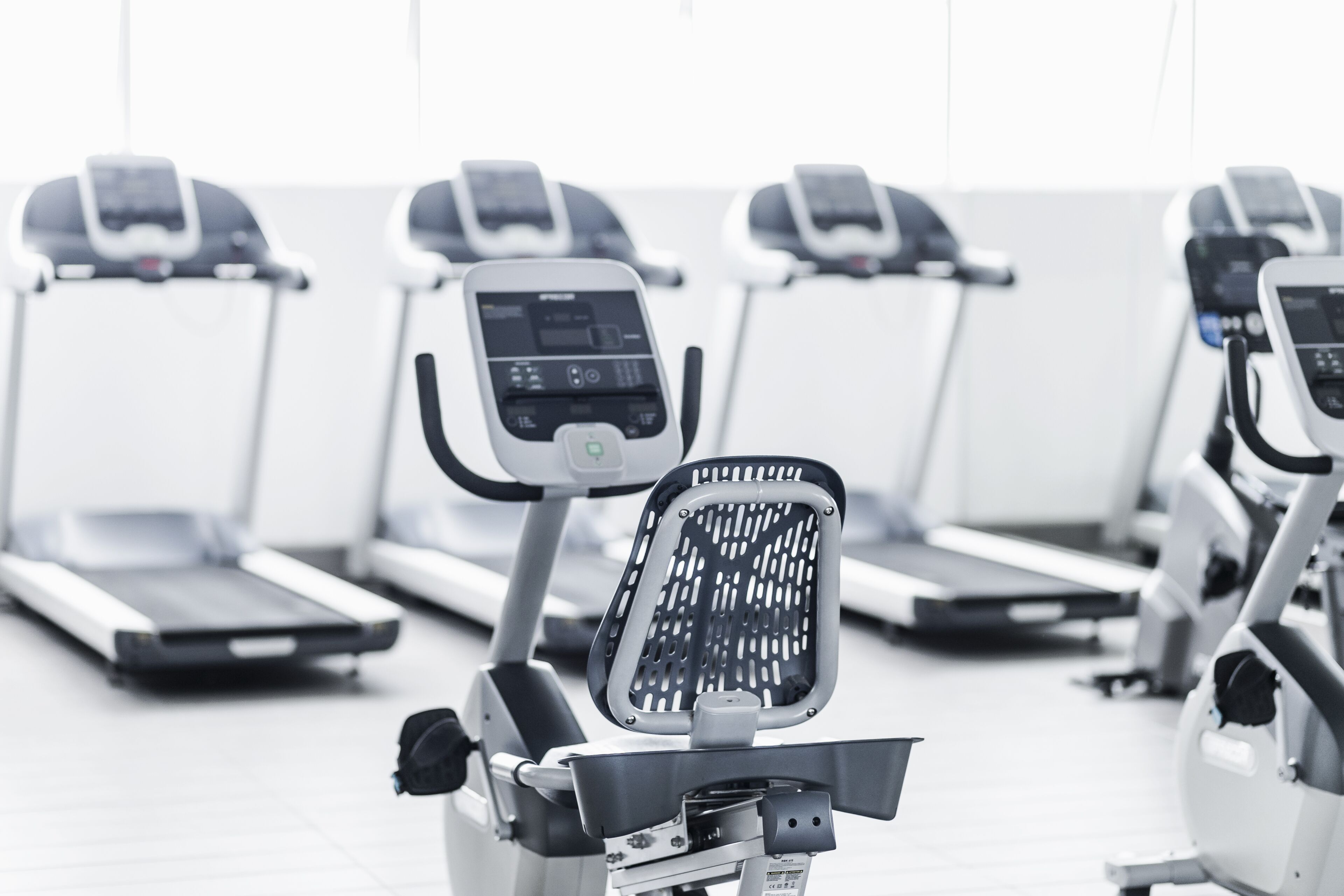 A row of stationary bikes in a gym with a white floor and a window in the background.