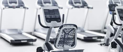 A row of stationary bikes in a gym with a white floor and a window in the background.