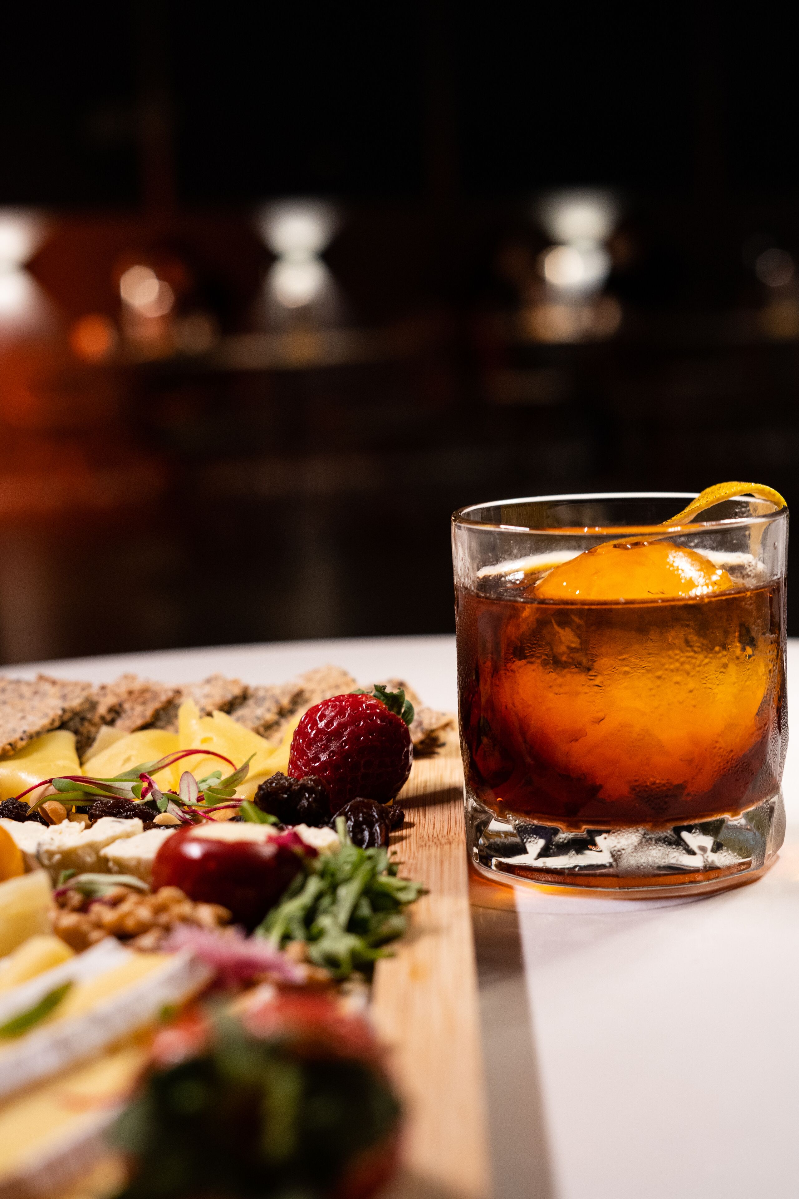 A glass of amber-colored beverage with a lemon slice, accompanied by an assortment of cheeses, fruits, and nuts on a wooden board.