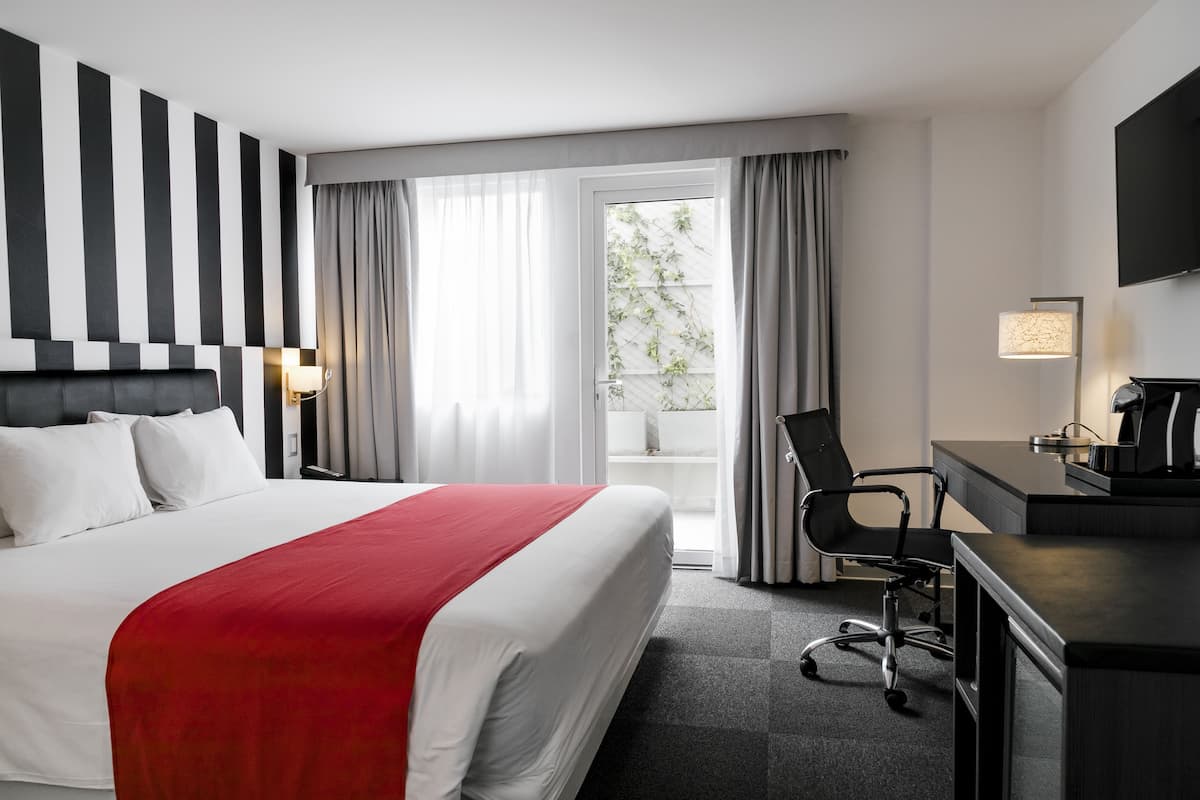 Habitación de hotel con cama, escritorio, silla, televisor y ventana con cortinas.
