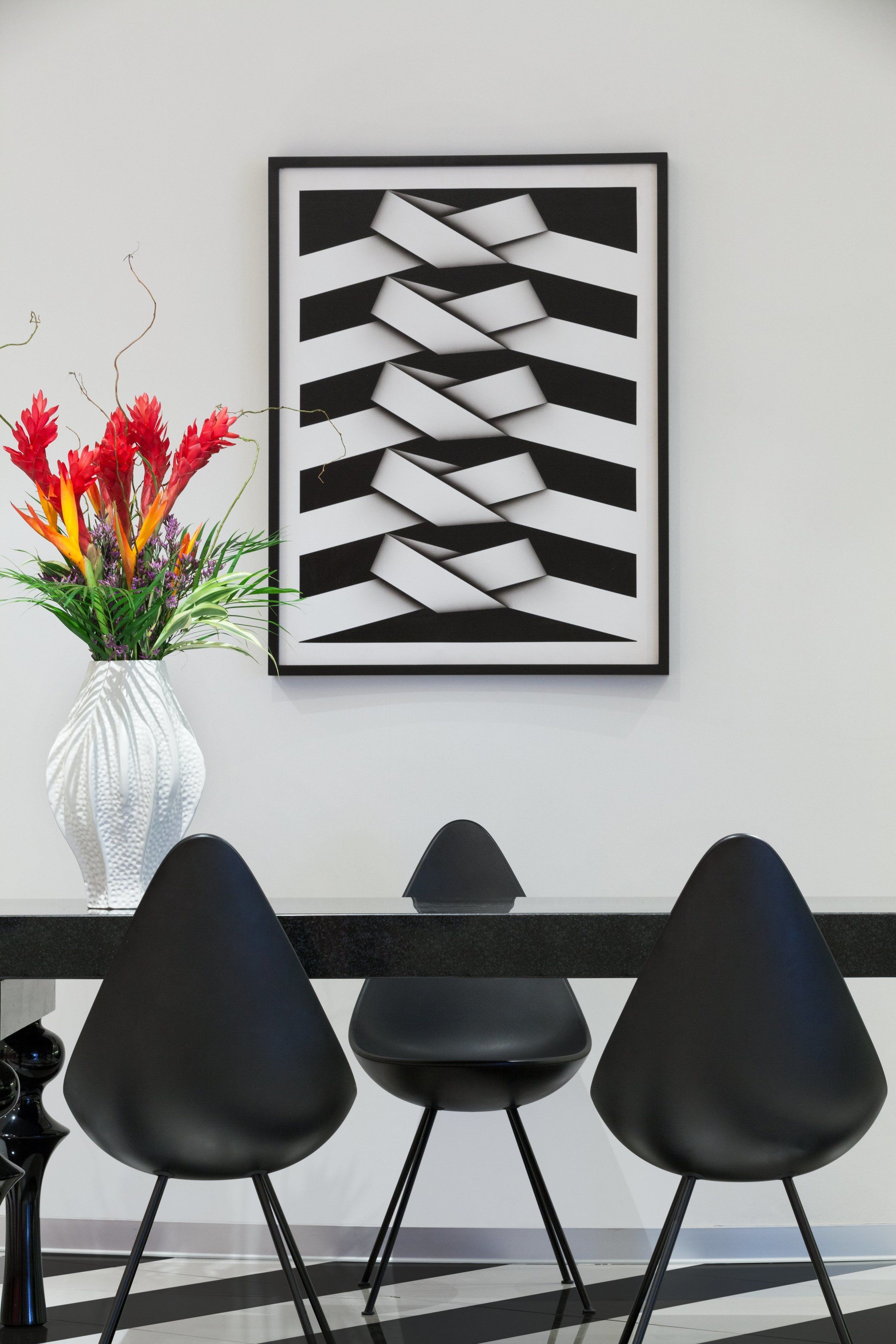 Un interior moderno con una pared blanca, una obra de arte abstracta enmarcada, un jarrón blanco con flores rojas y tres sillas negras.