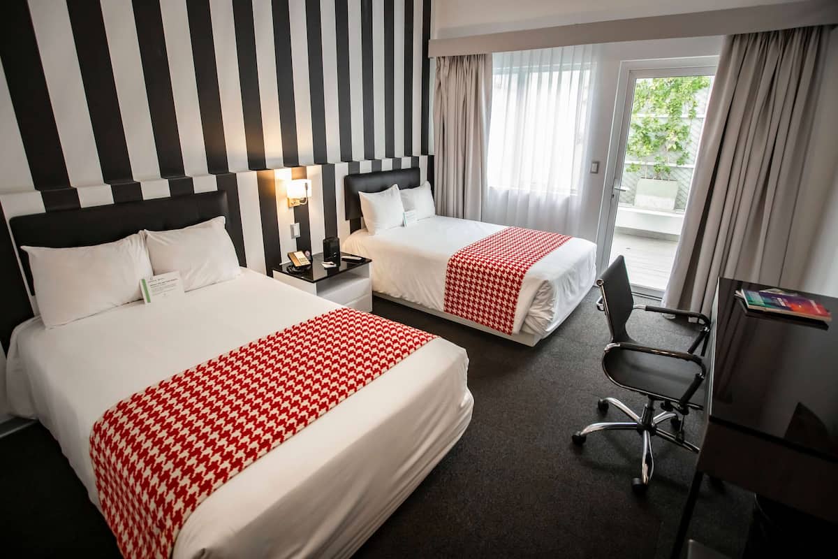 Habitación de hotel con dos camas, un escritorio y una silla. Tiene una ventana con cortinas y vistas a zonas verdes.