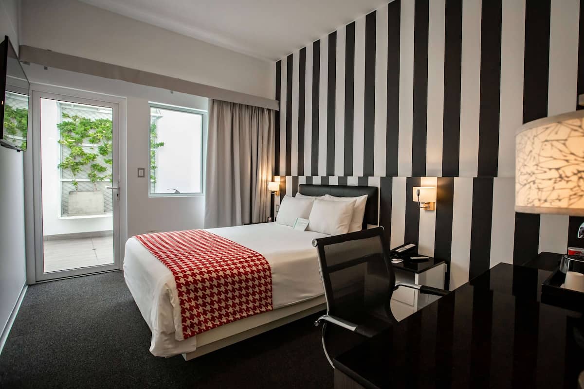 Habitación de hotel con cama, escritorio, silla, lámpara y vistas al exterior.