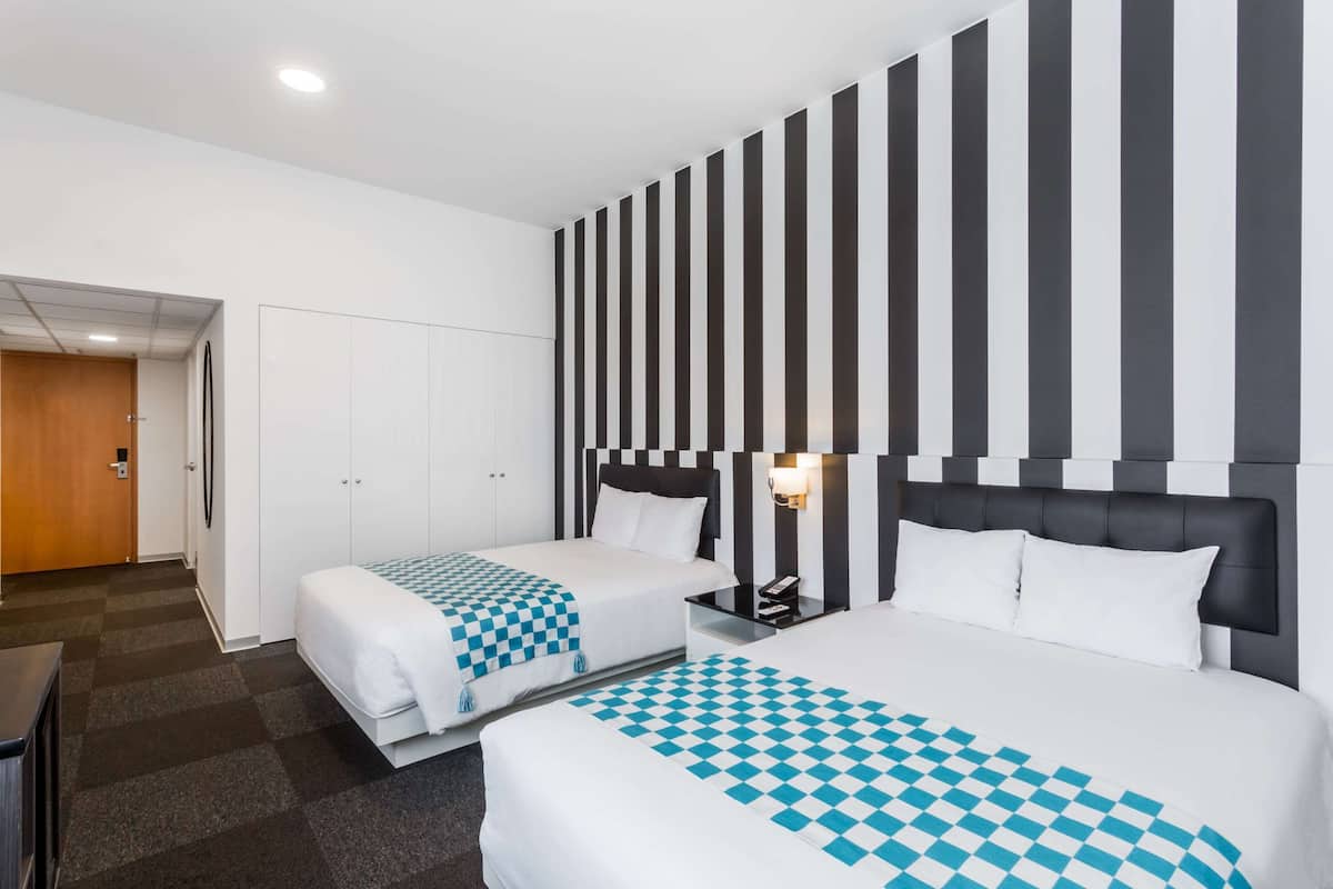 Habitación de hotel con dos camas, una colcha a cuadros, una pared a rayas y una mesita de noche con lámpara.