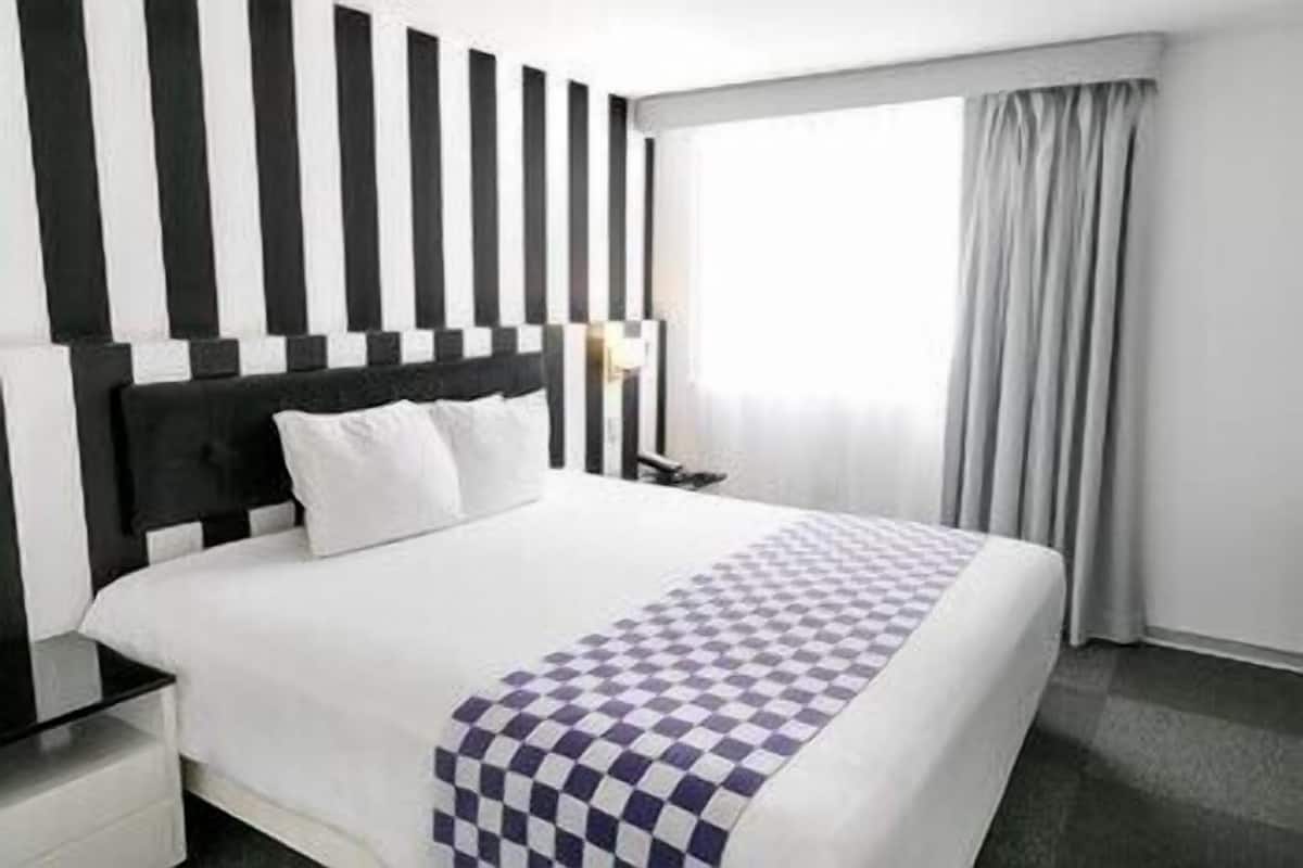 Habitación de hotel con una cama grande, cabecera con rayas verticales, manta a cuadros, mesita de noche y ventana con cortinas.
