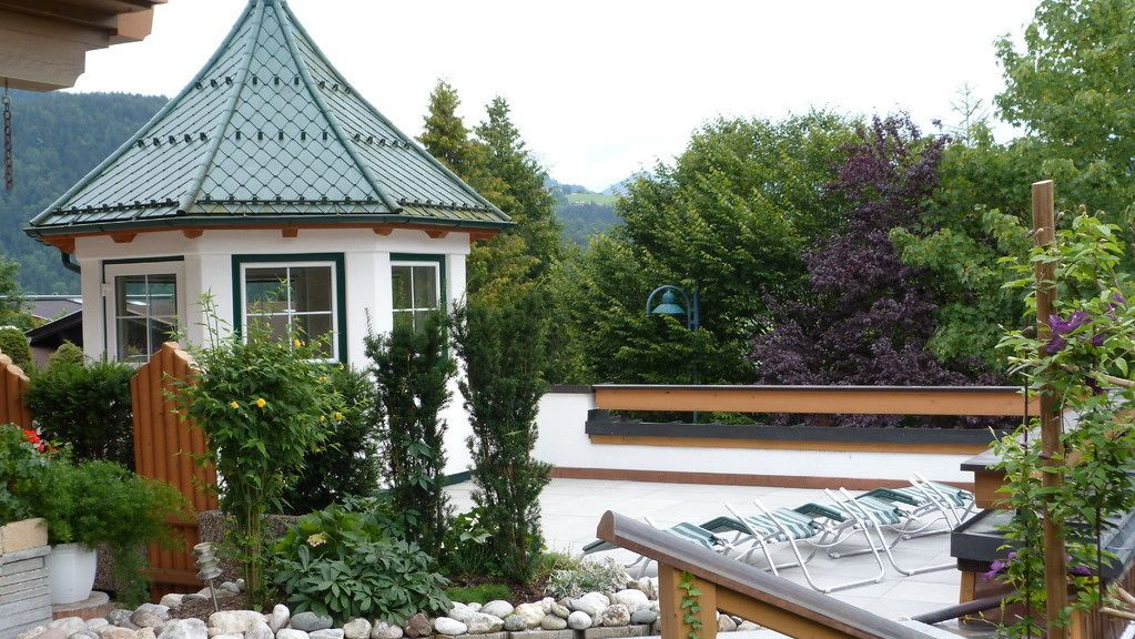 A gazebo with a green roof, surrounded by a garden with various plants and a wooden deck with chairs.