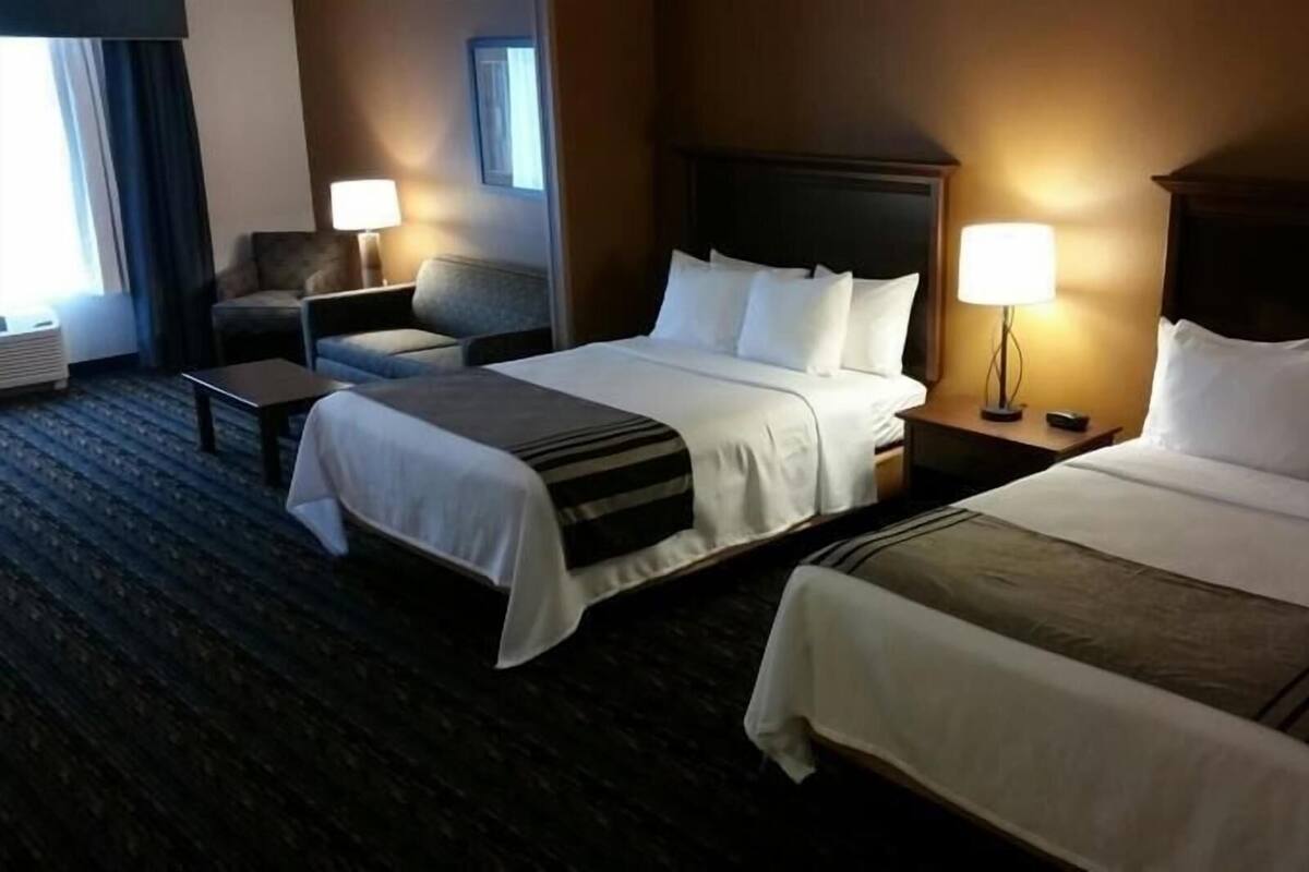 Quarto de hotel com duas camas, uma cadeira, uma mesinha e um abajur.