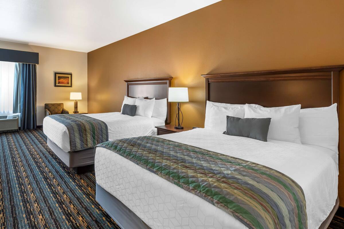 A hotel room with two beds, a large window with curtains, a wall-mounted lamp, and a patterned carpet.
